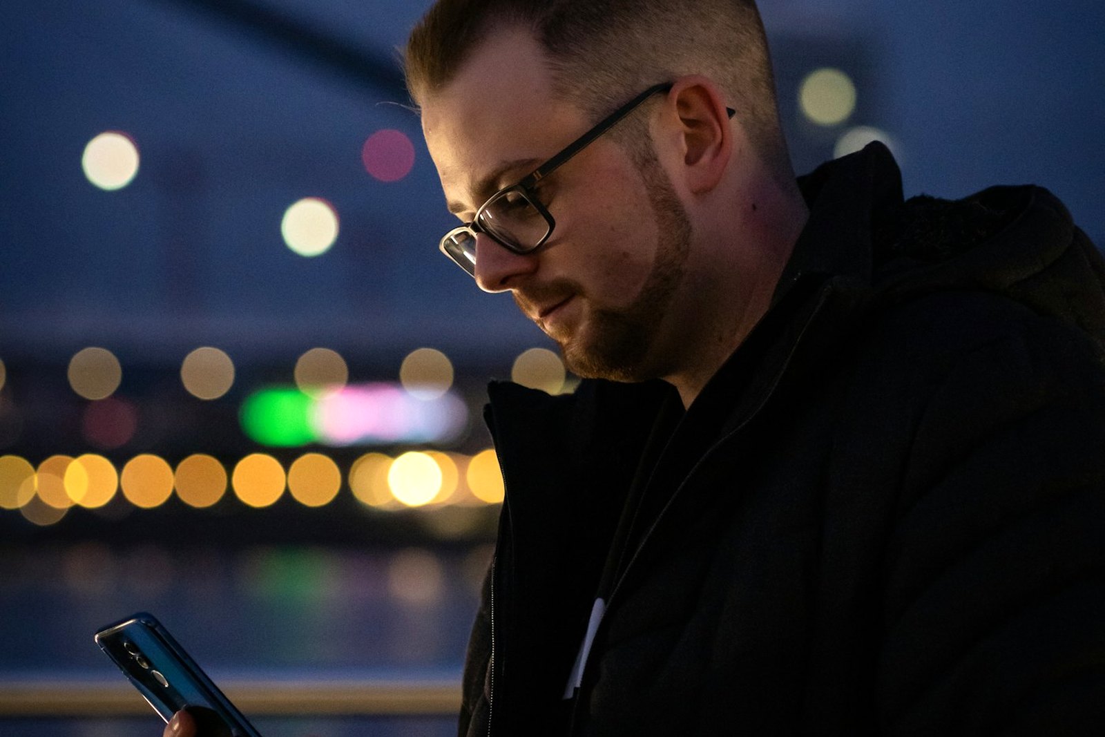 A man with glasses at night, looking down at the phone in his hand, city lights blurred behind him