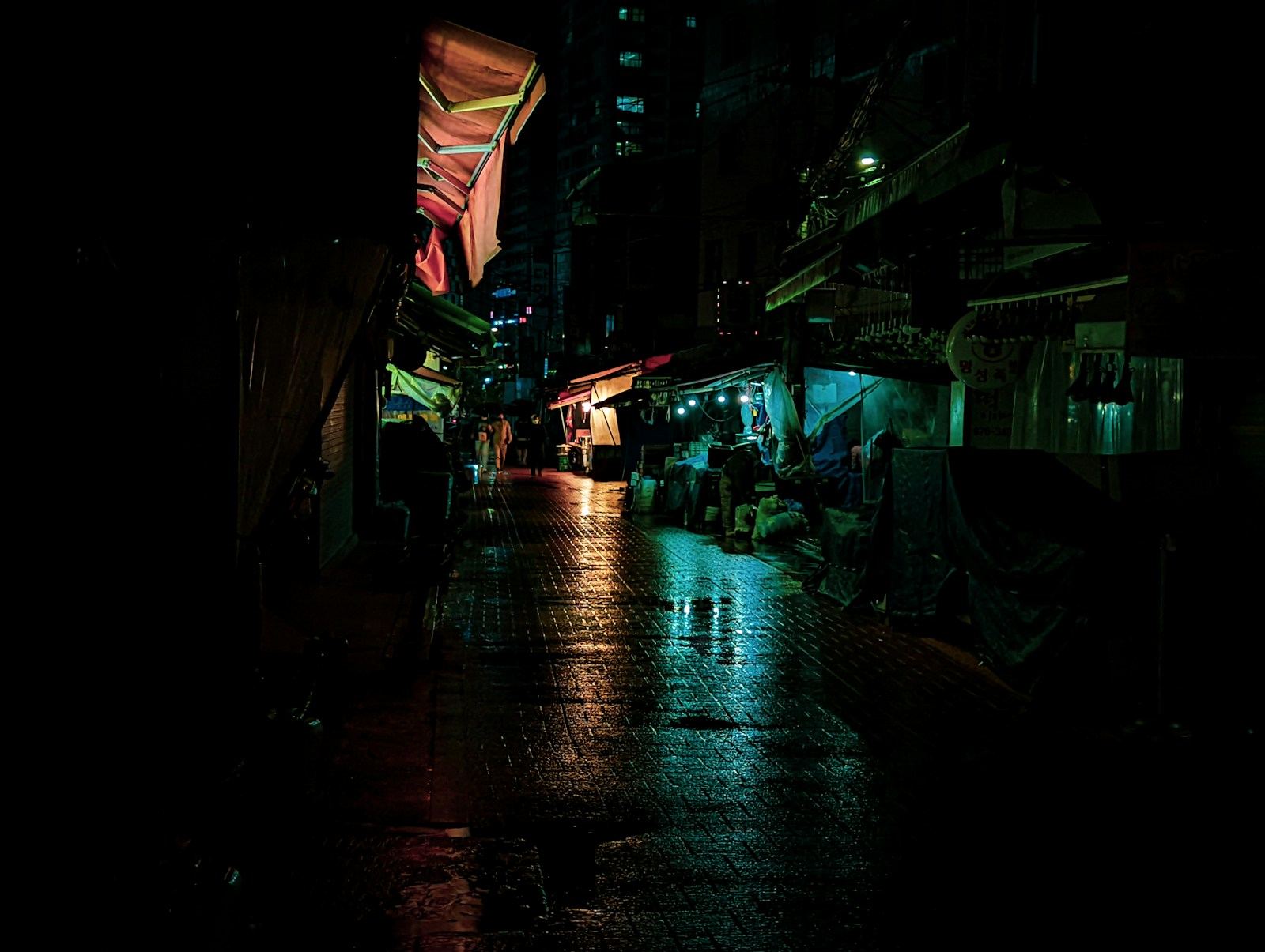 A narrow city street at night, wet pavement reflecting shop lights