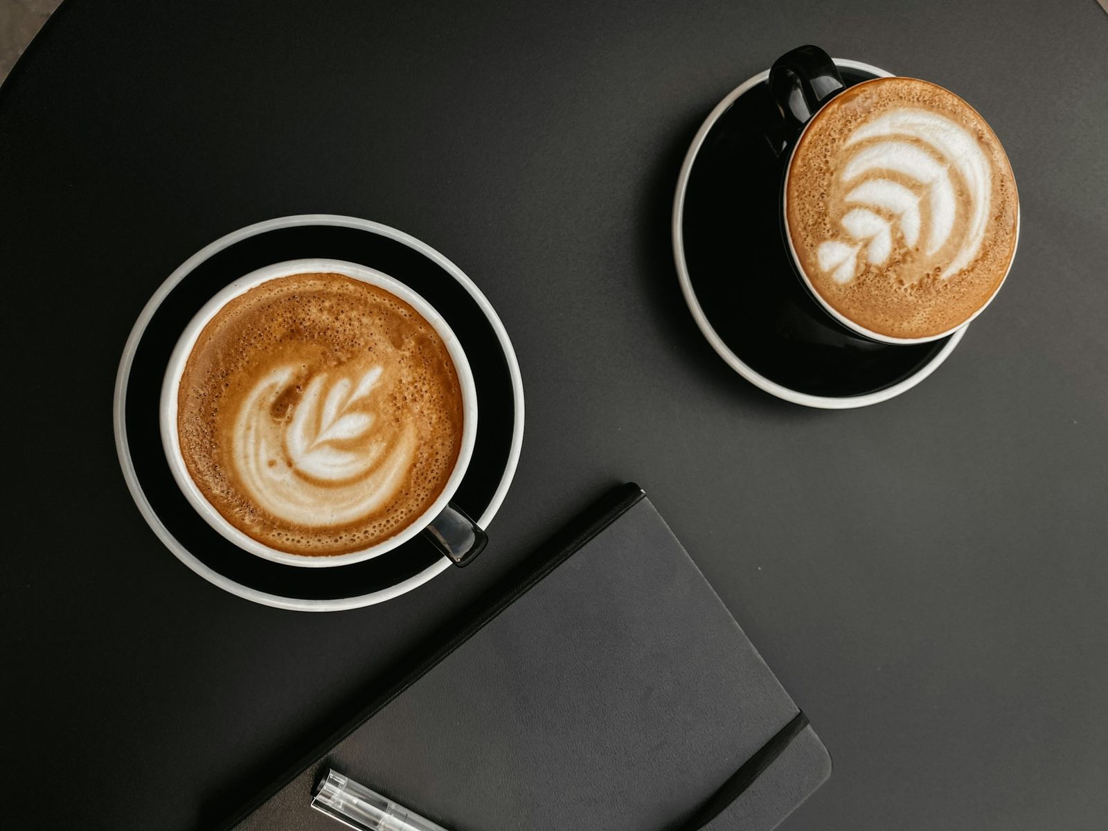 Two coffees and a closed black notebook on a dark table, seen from above