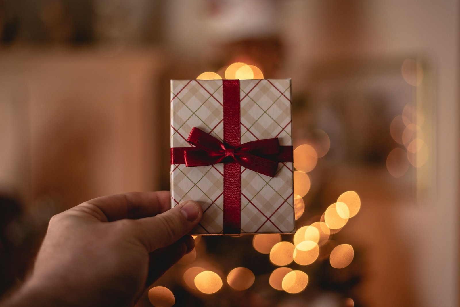 A man's hand holding up a small wrapped gift with a red ribbon, warm lights glowing behind it